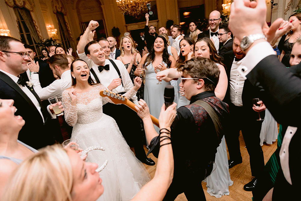 The guitarist from the band is playing a solo on the dance floor while the bride and groom cheer.