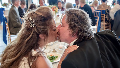 A wedding couple kiss at their table while guests applaud