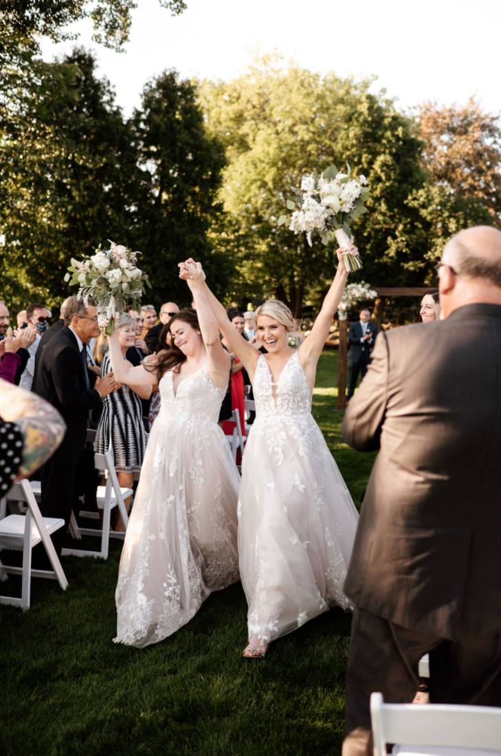 Two Brides walking down the aisle while their guests applaud.